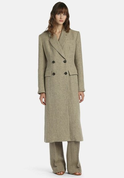 Femme portant un long manteau beige à double boutonnage avec des boutons noirs, un pantalon large assorti et des sandales ouvertes sur un fond blanc.