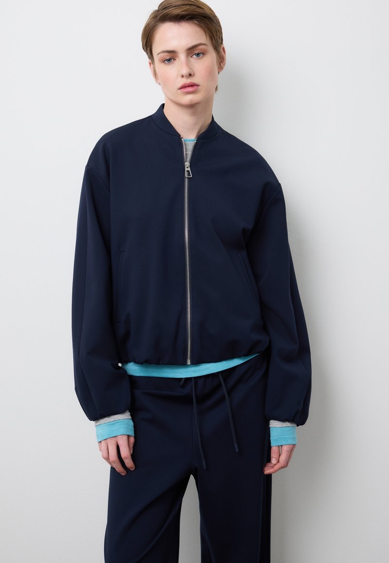 Marineblauer Bomberjacke mit Reißverschluss, elastischen Bündchen und einem türkisfarbenen Akzent am Saum. Kombiniert mit passenden marineblauen Hosen. Glatte Stofftextur.