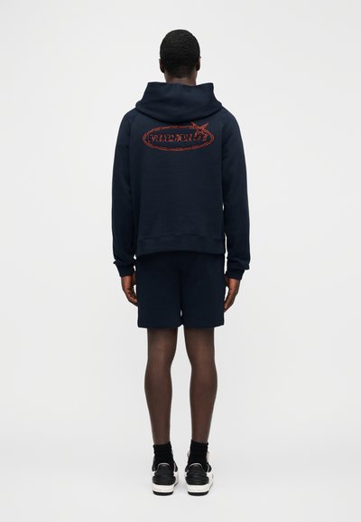 Námořnická mikina s kapucí s červeně vyšitým logem "VOLTAIRE", doplněná o odpovídající šortky, které ukazují volný střih a žebrované lemy.