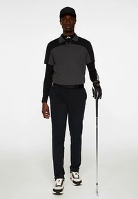 Golfkläder består av en grå piké med korta ärmar och svarta detaljer, kombinerad med marinblå byxor och svarta skor. En golfklubba hålls i ena handen.