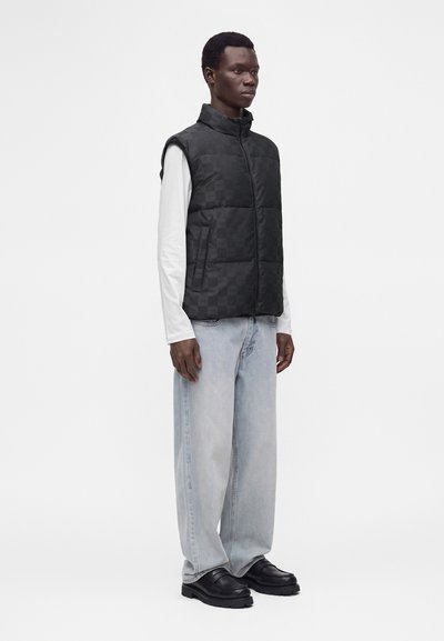 Homme portant un gilet matelassé à carreaux noir, une chemise blanche à manches longues, un jean large bleu clair, et des chaussures en cuir noires, debout de profil.