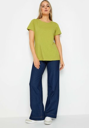 Camiseta verde de manga corta con cuello redondo, combinada con jeans de pierna ancha de color azul oscuro. Zapatillas grises y blancas completan el conjunto.