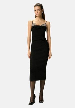 Žena v čiernych midi šatách bez rukávov s ozdobnou vrchnou časťou, čiernych priehľadných pančuchách a čiernych vysokých podpätkoch stojaca pred jednoduchým pozadím.