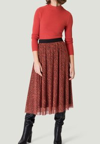 Top à manches longues rouge côtelé associé à une jupe plissée noire et rouge à motifs, complété par des bottes noires à talons montantes.