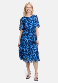 Niebieska sukienka w plisy z kwiecistym wzorem, przezroczystymi rękawami i podkreśloną talią. Połączona z niebieskimi sandałami na obcasie. Gładka tekstura, zwiewny krój.