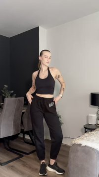 Black cropped halter top paired with black sweatpants featuring a white "GAP" logo. Model wears black sneakers, standing in a modern interior.