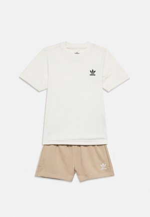Fehér Adidas póló fekete logóval a mellkason, bézs Adidas rövidnadrággal, amelynek bal lábán fehér logó található, fehér háttéren laposan elrendezve.