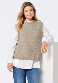 Beige gestrickter ärmelloser Pullover mit hohem Kragen, über einem weißen Hemd mit Kragen getragen, kombiniert mit blauen Jeans. Weiche Textur.
