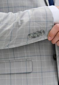 Grå ternet blazer med blå linjer, tekstureret stof, fire grå knapper på ærmet og en paspoleret lomme synlig på ærmet.