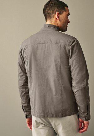 Veste légère grise avec un col montant, des coutures visibles et des poignets boutonnés. Le tissu semble lisse avec une finition légèrement texturée.