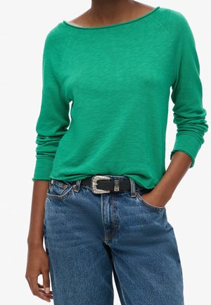 Osoba w zielonej bluzce z długim rękawem, wpuszczonej w niebieskie jeansy z wysokim stanem, z czarnym paskiem z srebrną klamrą.