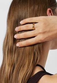 Dłoń z wypolerowanymi paznokciami i złotym pierścionkiem spoczywa na długich brązowych włosach, widoczne ramię i ucho, nosi ciemny top bez rękawów.