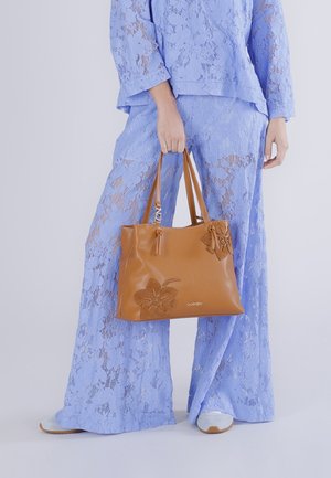 Persona che indossa un abito in pizzo azzurro chiaro e tiene una borsa in pelle marrone con ricamo floreale, in piedi davanti a uno sfondo neutro.