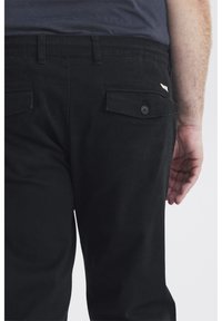 Pantalons noirs à texture lisse, dotés de deux poches arrière avec des fermetures à bouton et d'une petite étiquette sur le côté.