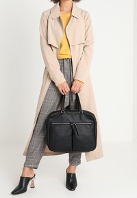 Kobieta w beżowym płaszczu przeciwdeszczowym, żółtym swetrze, kraciastych spodniach i czarnych szpilkach trzymająca dużą czarną skórzaną torbę za uchwyty.