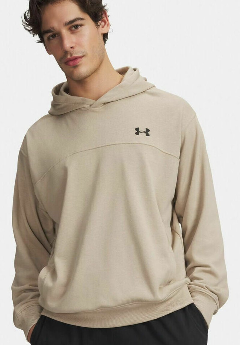Joven con sudadera con capucha beige de Under Armour, con las manos en los bolsillos, sobre fondo blanco.