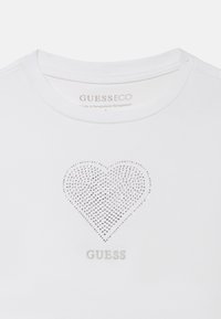 Hvid bomulds-T-shirt med et hjerte lavet af sølvrhinsten, med teksten "GUESS" nedenunder, rund halsudskæring og korte ærmer.