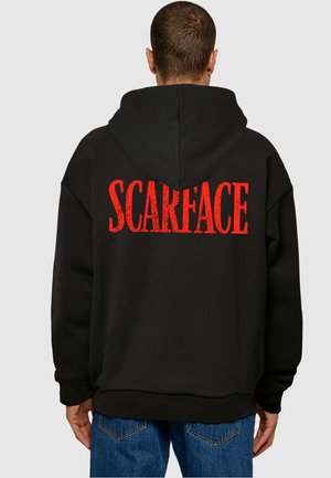 Man wearing black hoodie with red "SCARFACE" text on the back, paired with blue jeans, standing facing away against a plain background.