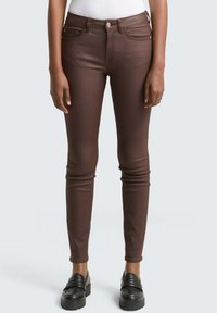 Oseba, oblečena v oprijete temno rjave hlače in črne �čevlje brez vezalk, stoji z rokami sproščeno ob straneh pred enostavno ozadje.