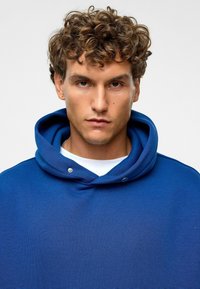 Blauwe hoodie met een grote capuchon, gemaakt van zacht materiaal, met drukknopen bij de hals en een ongestructureerd silhouet.