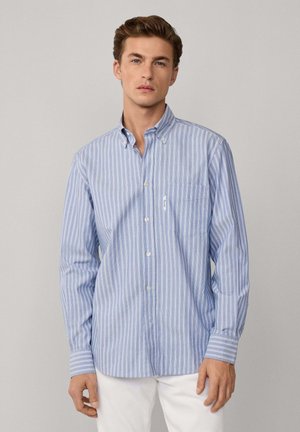 Hombre vestido con una camisa de botones a rayas azul claro y blanca con bolsillo en el pecho y pantalones blancos, de pie frente a un fondo gris liso.