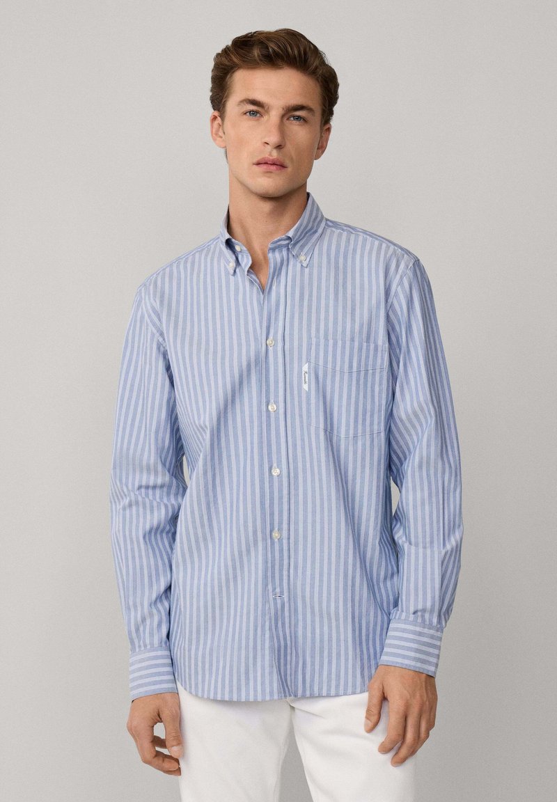 Hombre vestido con una camisa de botones a rayas azul claro y blanca con bolsillo en el pecho y pantalones blancos, de pie frente a un fondo gris liso.