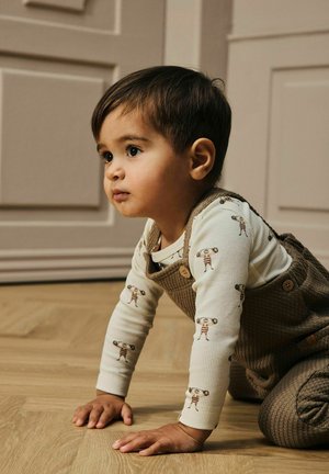 Un jeune enfant aux cheveux foncés qui rampe sur un sol en bois, portant une salopette marron et une chemise crème avec un imprimé de haltérophile, regardant intensément devant lui.