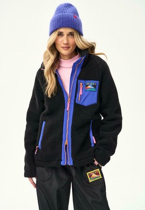 Kobieta ubrana w czarną polarową kurtkę z niebieskimi i różowymi zamkami, dopasowaną niebieską czapkę, różowy golf oraz czarne spodnie z kolorowymi naszywkami "Explorers".