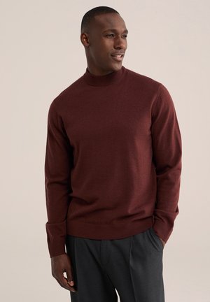 Homme portant un pull à manches longues bordeaux et un pantalon gris foncé, debout avec une main dans la poche, regardant sur le côté devant un fond uni.