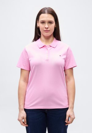 Woman with long brown hair wearing gold hoop earrings, a pink short-sleeve polo shirt, and dark blue jeans, facing forward against a plain background.