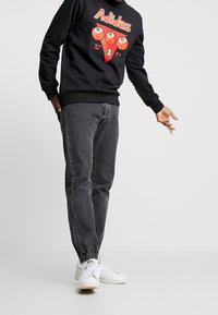 Person wearing black sweatshirt with red tomato graphic and Adidas text, dark gray jogger jeans, and white sneakers, extending one arm sideways.