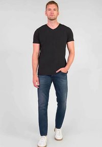 Svart kortärmad V-ringad T-shirt kombinerad med tajta blå jeans och vita sneakers, mot en enfärgad grå bakgrund.