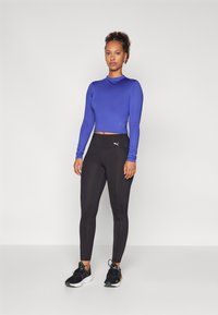 Blue long-sleeve athletic top with a fitted design, paired with black high-waisted leggings. Model wearing black sneakers with colorful accents.