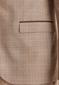 Gros plan d'une poche de blazer beige et marron à carreaux avec un rabat et une bordure marron foncé contrastante.