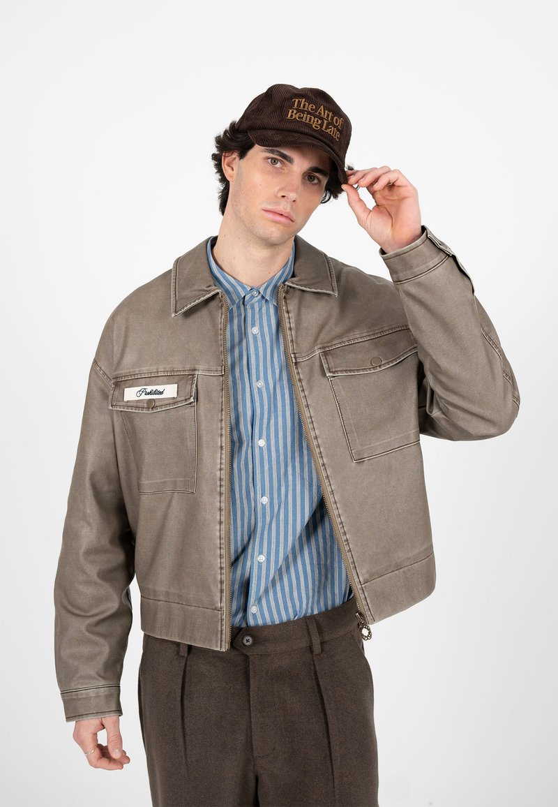 Jeune homme ajustant une casquette marron foncé avec l'inscription « The Art of Being Late », portant une veste beige sur une chemise rayée bleue et un pantalon marron.