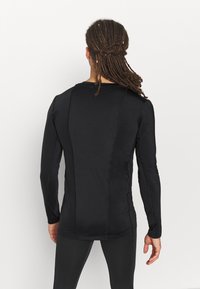 Ein Mann mit langen Dreadlocks, der ein enges schwarzes Langarmhemd und schwarze Hosen trägt, steht vor einem schlichten weißen Hintergrund.