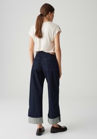 Jeans a gamba larga blu scuro con risvolti, abbinati a una maglietta corta crema, caratterizzata da un design semplice e un minimo di dettagli.