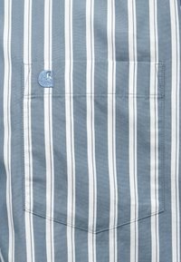 Svetlomodrá košeľa s vertikálnymi bielymi prúžkami. Obsahuje malú náprsné vrecko a vyšívané logo v modrej farbe. Mäkký bavlnený materiál.