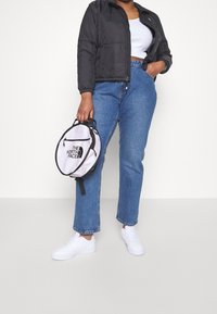 Svart vadderad jacka, vit kort topp, högmidjade blå jeans och vita sneakers. Håller en vit och svart rund ryggsäck med logotyp.