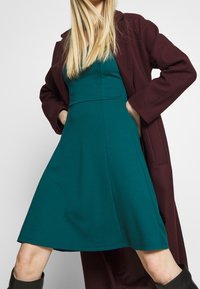 Robe bleu sarcelle avec une jupe évasée et un corsage ajusté, superposée sous un manteau bordeaux. Tissu doux, manches longues et poches latérales visibles.