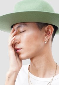 Young person wearing a light green hat, with closed eyes, touching their face, and wearing multiple earrings and layered gold necklaces.