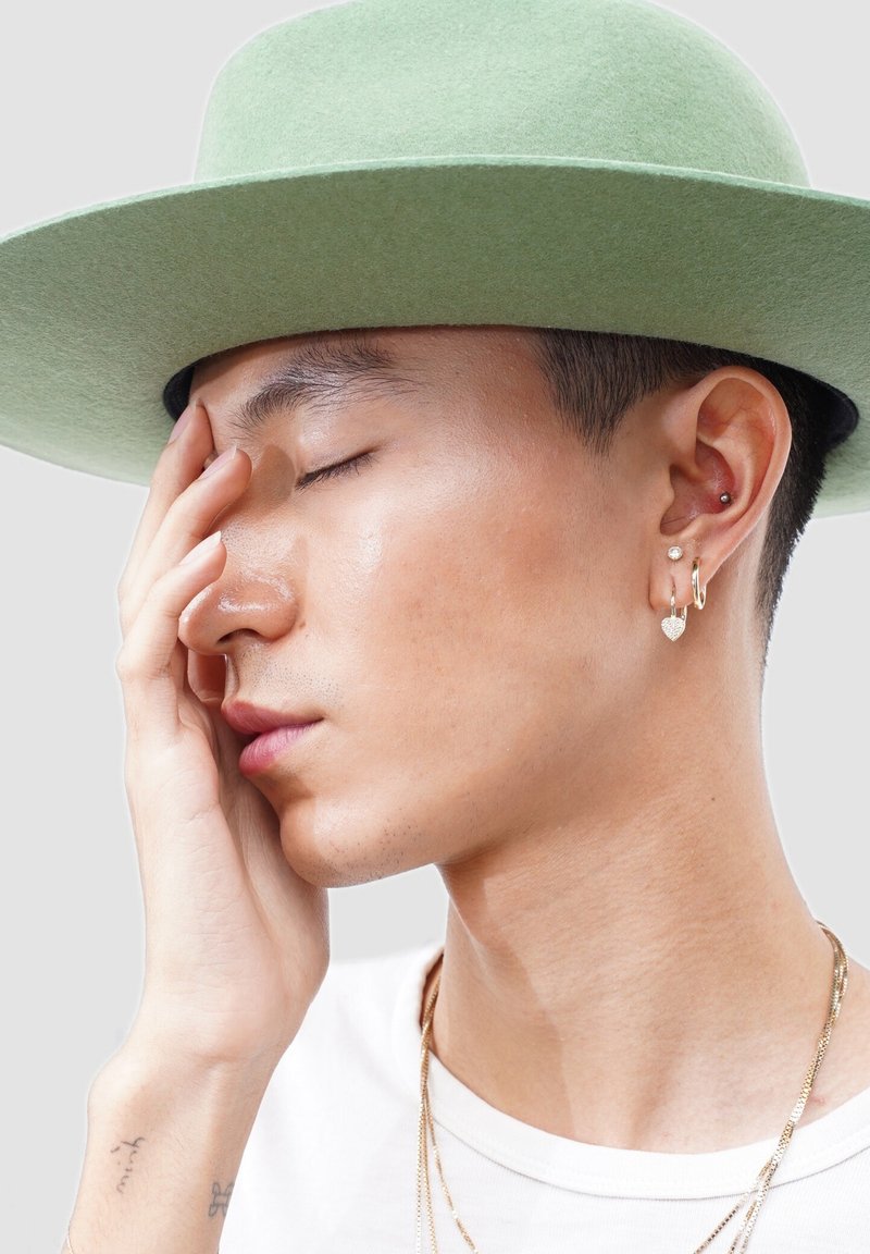 Young person wearing a light green hat, with closed eyes, touching their face, and wearing multiple earrings and layered gold necklaces.