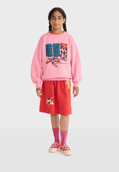 Jeune fille aux cheveux tressés portant un sweat-shirt rose avec un motif de chien, une jupe rouge ornée de motifs de vache et de personnage, des chaussettes rayées et des chaussures rouges.