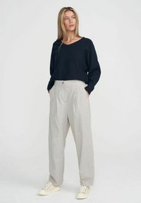 Marineblauer V-Ausschnitt-Pullover kombiniert mit grau gestreiften, hoch taillierten Hosen und cremefarbenen Sneakers. Das Model steht mit den Händen in den Taschen.
