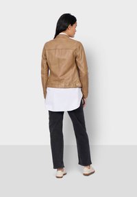 Veste en cuir tan avec une coupe ajustée, coutures au dos et côtés boutonnés, superposée à une longue chemise blanche et associée à un pantalon et des chaussures foncés.