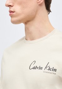 Κοντινό πλάνο του κάτω μέρους του προσώπου και του λαιμού ενός ατόμου που φοράει ένα ανοιχτό μπεζ πουκάμισο με την επιγραφή «Calvin Klein Established 1968» στο στήθος.