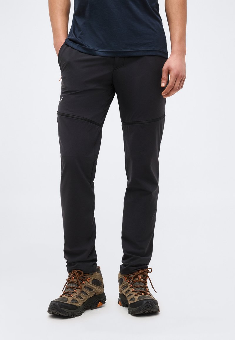 Άτομο που φοράει μαύρο παντελόνι για πεζοπορία με φερμουάρ και καφέ παπούτσια πεζοπορίας, όρθιο με τα χέρια χαλαρά στο πλάι.