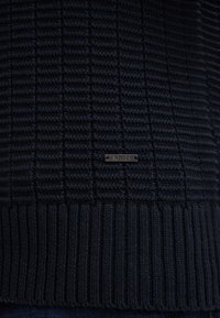 Dunkelblauer Strickpullover mit strukturiertem Muster, geripptem Saum und einem kleinen metallischen Logo-Tag mit "Joop!" auf der Vorderseite.