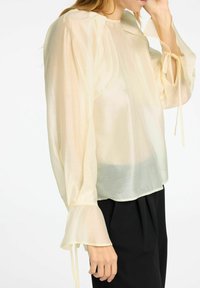 Blusa leggera color beige chiaro, trasparente, con maniche lunghe e svasate e dettagli legati ai polsi. Tessuto morbido e liscio con una vestibilità rilassata.