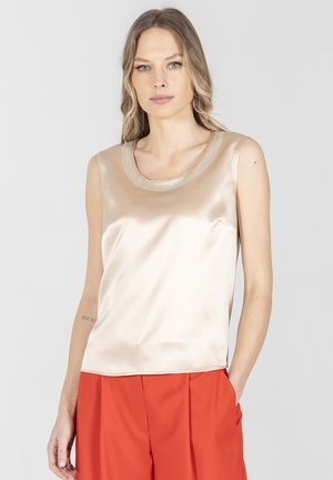 Kobieta o długich blond włosach, ubrana w beżowy satynowy top bez rękawów z kryształkami na dekolcie oraz czerwone spodnie, stojąca z jedną ręką w kieszeni.